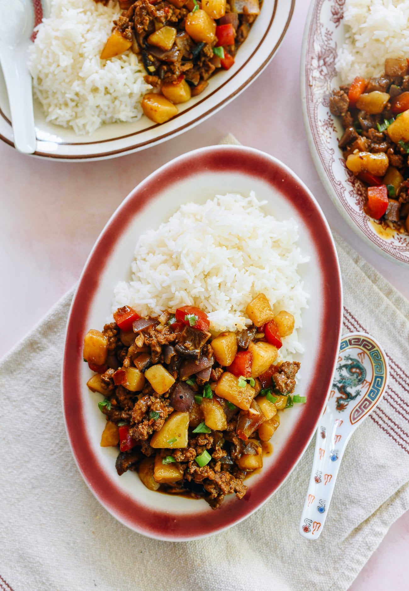 Chines braised pork, potato, and bell pepper over rice on a red rimmed oval plate on a pink marble counter