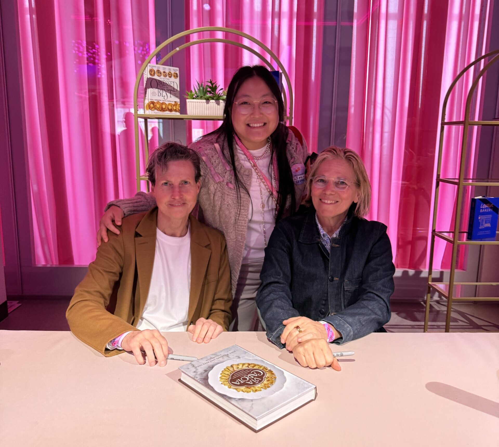 Kaitlin Leung at a book signing with Via Carota chefs Rita Sodi and Jody Williams at a pink table in front of a hot pink wall.